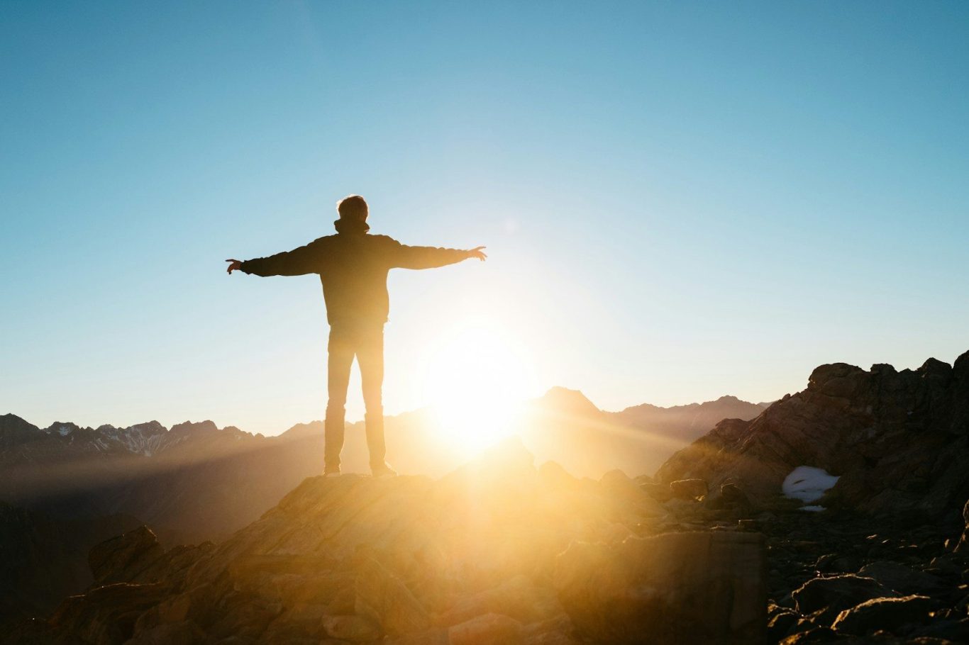 Bild: Eine Person steht auf einem Berg, streckt die Arme zu den Seiten aus. Die Sonne geht unter und taucht das Bild in helles Licht
