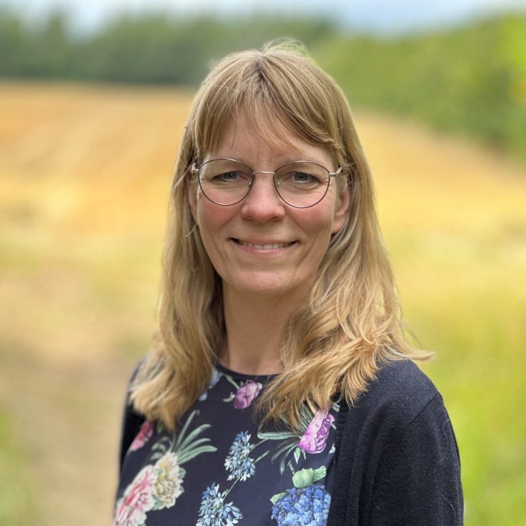 Bild: Ein Bild von Iris Kruck. Sie hat lange blonde Haare, trägt eine rote Jacke und im Hintergrund ist das Meer zu sehen.