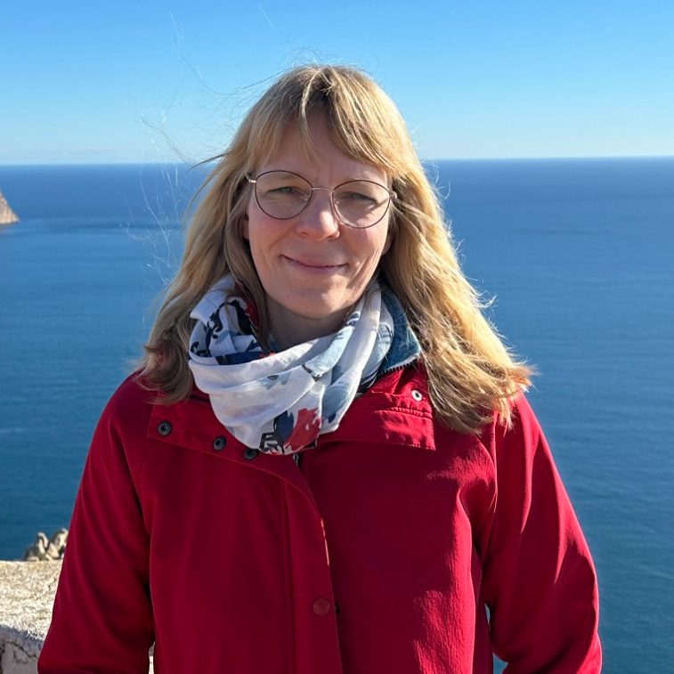 Bild: Ein Bild von Iris Kruck. Sie hat lange blonde Haare, trägt eine rote Jacke und im Hintergrund ist das Meer zu sehen.